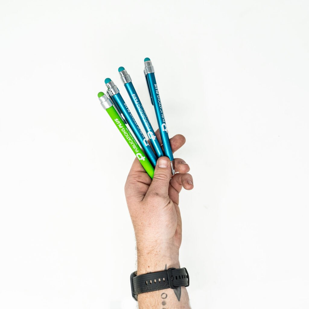 Hand holding multiple green and blue pens with Publications Plus logo on it.